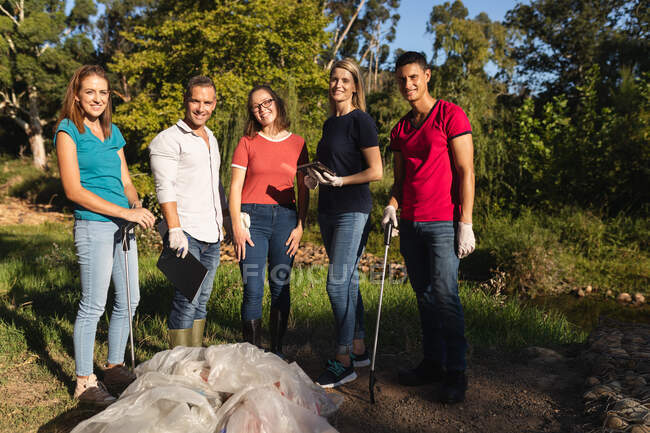 Ritratto di felice gruppo multietnico di volontari per la conservazione che ripuliscono il fiume in campagna, sacchi di rifiuti raccolti di fronte. Ecologia e responsabilità sociale nell'ambiente rurale. — Foto stock