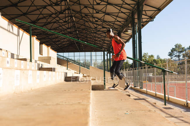 In forma, atleta maschio disabile di razza mista in uno stadio sportivo all'aperto, salendo le scale sugli spalti indossando cuffie e lame da corsa. Disabilità atletica allenamento sportivo. — Foto stock