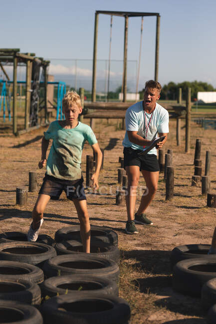 Кавказский мальчик в лагере в солнечный день, в грязной зеленой футболке и черных шортах, бегающий по шинам на полосе препятствий, с кавказским тренером, кричащим на заднем плане — стоковое фото