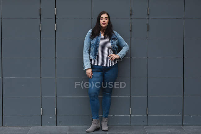 Portrait de femme caucasienne courbée dans les rues de la ville pendant la journée, debout avec sa main sur la hanche regardant vers la caméra, appuyé contre un mur gris foncé — Photo de stock