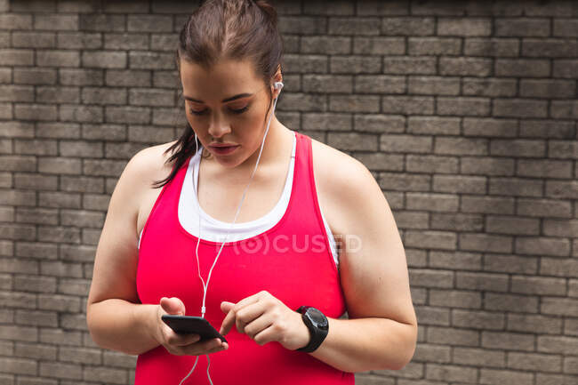Mujer curvilínea caucásica con el pelo largo y oscuro usando ropa deportiva haciendo ejercicio en una ciudad, usando su teléfono inteligente con auriculares, una pared de ladrillo en el fondo - foto de stock
