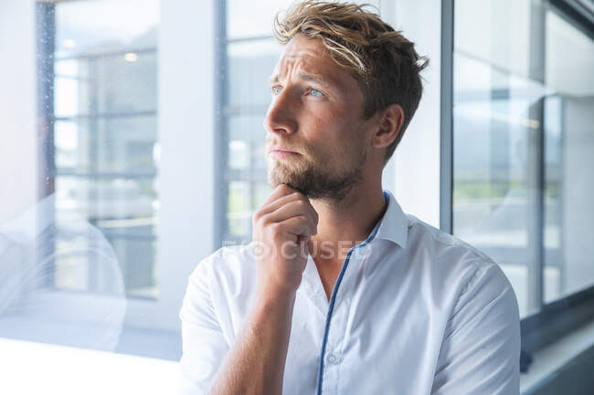 Smart casually dressed Caucasian male business creative with blonde hair and beard looking out of window thinking, with hand on his chin. Creative business professional working in a modern office. — Stock Photo