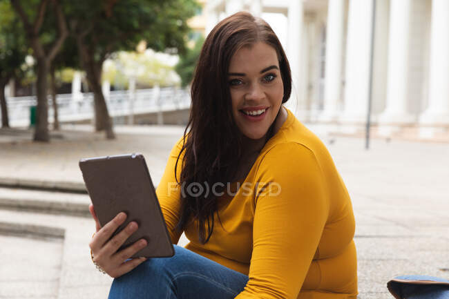 Retrato de una mujer caucásica con curvas por las calles de la ciudad durante el día, sentada en escalones usando una tableta digital y sonriendo a la cámara, con un edificio histórico en el fondo - foto de stock