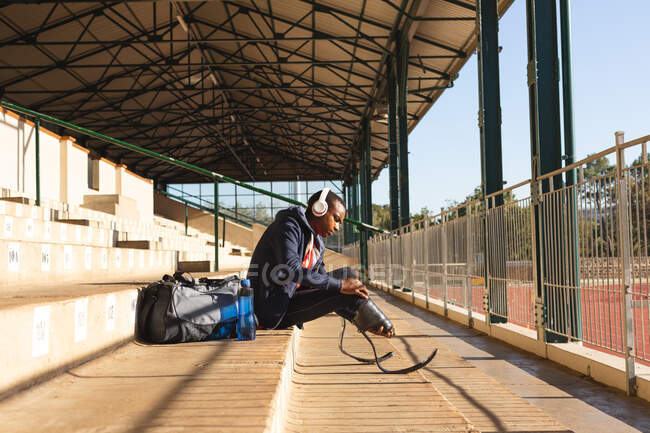 Ajustement, athlète masculin handicapé de race mixte dans un stade de sport de plein air, assis dans les gradins portant des écouteurs ajustant les lames de course. Handicap athlétisme entraînement sportif. — Photo de stock
