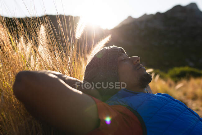 Подтянутый спортсмен смешанной расы, наслаждающийся поездкой в горы, отдыхающий на траве с закрытыми глазами и руками за головой. Активный образ жизни. — стоковое фото