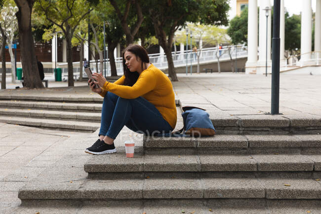 Mujer curvilínea caucásica por las calles de la ciudad durante el día, sentada en escalones con un café para llevar y su mochila, usando una tableta digital, con un edificio histórico en el fondo - foto de stock