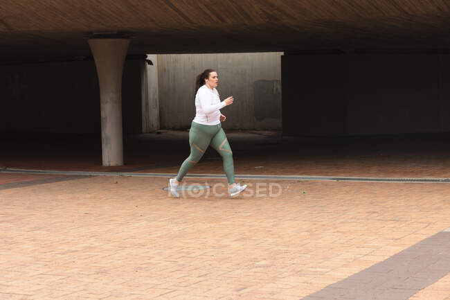 Mujer curvilínea caucásica con el pelo largo y oscuro usando ropa deportiva haciendo ejercicio en una ciudad, corriendo con auriculares en una zona peatonal urbana - foto de stock