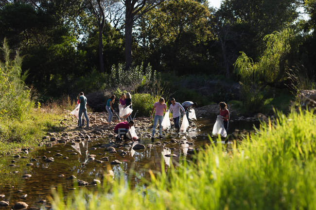 Un gruppo multietnico di volontari per la conservazione pulisce il fiume nelle campagne, raccogliendo spazzatura. Ecologia e responsabilità sociale nell'ambiente rurale. — Foto stock