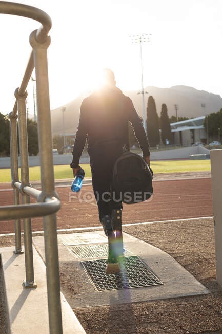 Rückansicht eines fitten, Mixed Race behinderten männlichen Athleten in einem Outdoor-Sportstadion, der mit Turnbeutel und Wasserflasche mit Laufschuhen läuft. Behindertensport. — Stockfoto