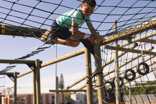 Niño caucásico en un campamento de entrenamiento en un día soleado, trepando a través de una red en un marco de escalada, usando una sudadera blanca, camiseta verde y pantalones cortos negros - foto de stock