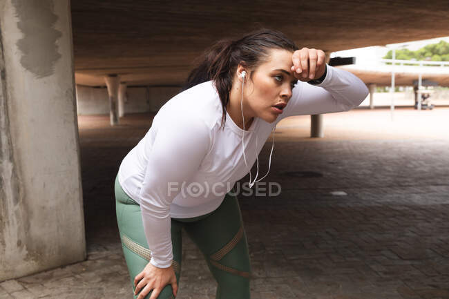 Кудрявая белая женщина с длинными темными волосами в спортивной одежде и наушниках, тренирующаяся в городе, отдыхающая и остывающая во время тренировки — стоковое фото