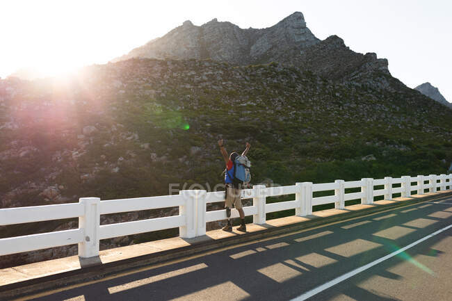Ein fitter, behinderter Mixed Race männlicher Athlet mit Beinprothese, der seine Zeit auf einem Ausflug in die Berge genießt und mit ausgestreckten Armen auf der Straße steht. Aktiver Lebensstil mit Behinderung. — Stockfoto