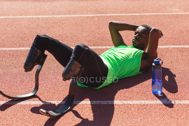 Fit, mixed race disabled male athlete at an outdoor sports stadium, lying on race track after race with water bottle wearing running blades. Disability athletics sport training. — Stock Photo