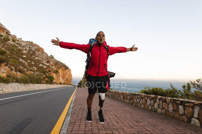 Un atleta masculino de raza mixta en forma y discapacitado con una pierna protésica, disfrutando de su tiempo en un viaje a las montañas, haciendo senderismo con los brazos extendidos en la carretera junto al mar. Estilo de vida activo con discapacidad. - foto de stock