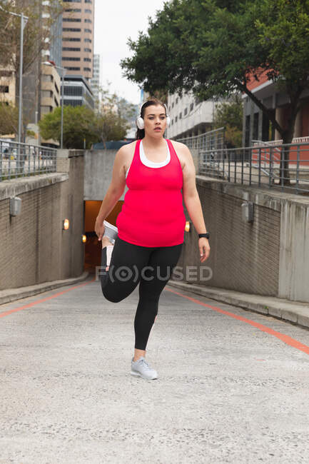 Curvy Caucasian woman with long dark hair wearing sports clothes exercising in a city with headphones on, holding her foot behind her and stretching her leg, with modern buildings in the background — Stock Photo