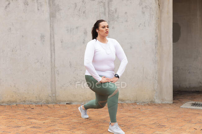 Кудрявая белая женщина с длинными темными волосами в спортивной одежде и наушниках, тренирующаяся в городе, растягивающаяся и разогревающаяся перед тренировкой — стоковое фото