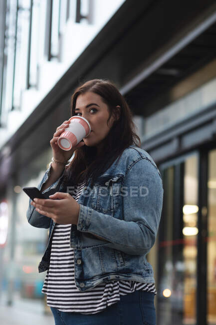 Curvy femme caucasienne dans les rues de la ville pendant la journée, boire un café à emporter et en utilisant son smartphone avec un bâtiment moderne en arrière-plan — Photo de stock