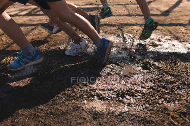 Seção baixa de um grupo de crianças em um acampamento de treinamento em um dia ensolarado, usando shorts e treinadores, correndo através da lama em um curso de obstáculo — Fotografia de Stock