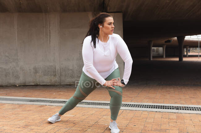 Mulher branca curvilínea com cabelos longos e escuros vestindo roupas esportivas e fones de ouvido exercitando-se em uma cidade, alongamento e aquecimento antes de seu treino — Fotografia de Stock