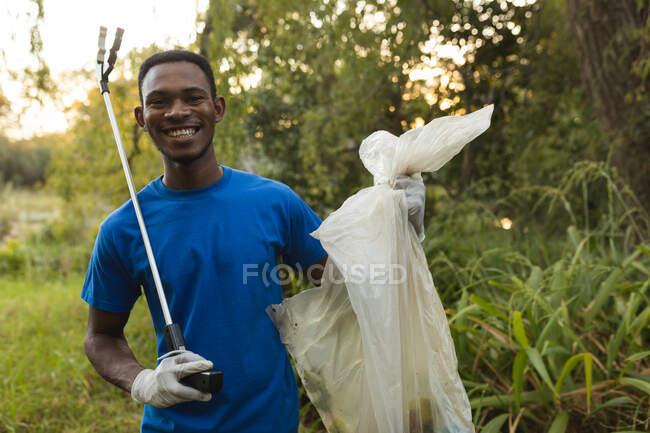 Retrato de un voluntario de conservación afroamericano limpiando el bosque en el campo, sonriendo sosteniendo la bolsa de basura y el agarrador. Ecología y responsabilidad social en el medio rural. - foto de stock