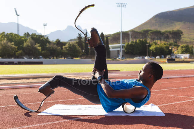 Fit, mixed race disabled male athlete at an outdoor sports stadium, preparing before workout stretching on roller on race track wearing running blades. Disability athletics sport training. — Stock Photo