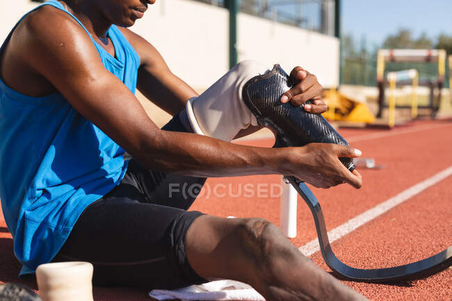 Mid section of fit, mixed race disabled male athlete at an outdoor sports stadium, sitting on race track preparing running blades before workout. Disability athletics sport training. — Stock Photo