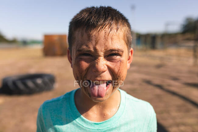 Retrato de niño caucásico con el pelo corto y oscuro y barro en la cara mirando a la cámara, sacando la lengua y haciendo una cara en un campamento de entrenamiento en un día soleado, vistiendo una camiseta verde - foto de stock