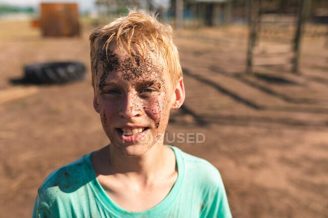 Portrait d'un garçon caucasien heureux avec des cheveux blonds courts dans un camp de démarrage par une journée ensoleillée, avec de la boue sur le visage portant un t-shirt vert sale, regardant la caméra et souriant — Photo de stock
