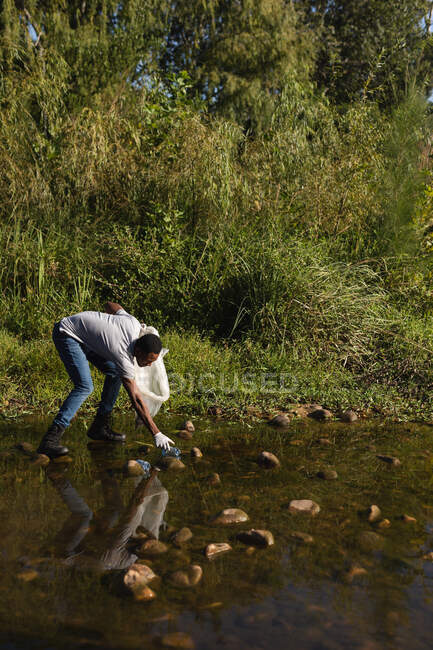 Hombre afroamericano voluntario de conservación limpiando el río en el campo, recogiendo basura. Ecología y responsabilidad social en el medio rural. - foto de stock