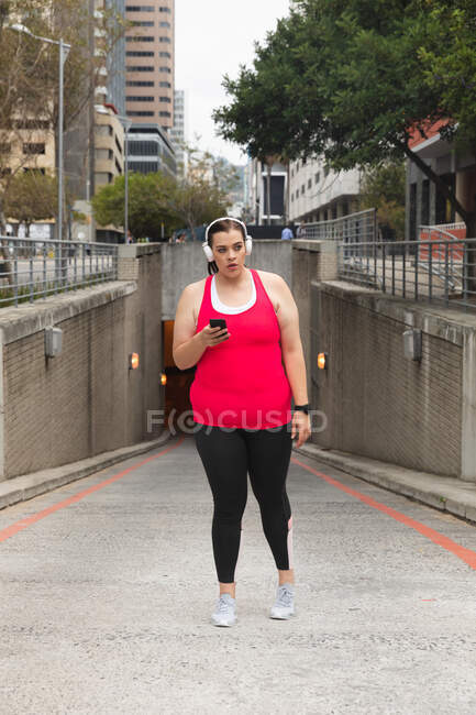 Curvy Caucasian woman with long dark hair wearing sports clothes exercising in a city, using her smartphone with headphones on, with modern buildings in the background — Stock Photo