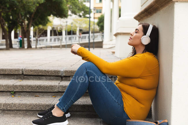 Femme caucasienne courbée dans les rues de la ville pendant la journée, assise sur des marches, penchée en arrière avec les yeux fermés et écoutant de la musique sur des écouteurs, avec un bâtiment historique en arrière-plan — Photo de stock