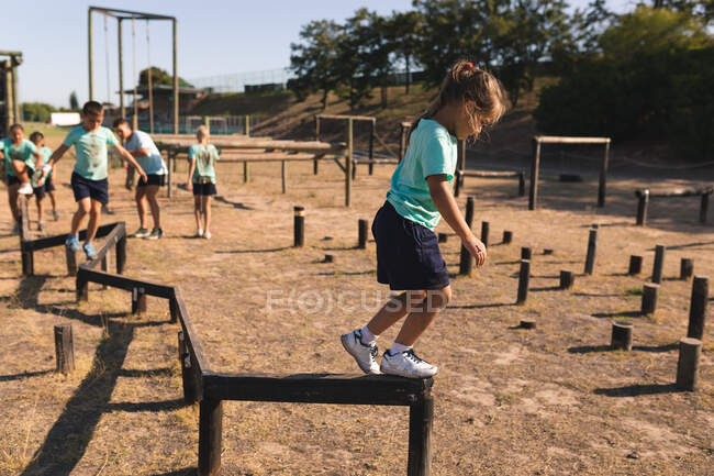 Кавказская девушка в лагере в солнечный день, в зеленой футболке и черных шортах, балансирует и идет вдоль бревна на полосе препятствий, а другие дети следуют за ней на заднем плане — стоковое фото