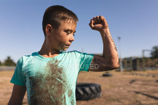 Кавказький хлопчик з коротким темним волоссям в чоботному таборі в сонячний день, з брудом на обличчі і руці і в брудній зеленій футболці, дивлячись на його руку і гнучкий біцепс — стокове фото