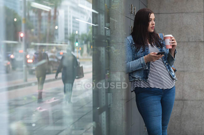 Kurvige Kaukasierin, die tagsüber in den Straßen der Stadt unterwegs ist, einen Kaffee zum Mitnehmen in der Hand hält und ihr Smartphone an ein modernes Gebäude lehnt, in dem sich die Straße spiegelt — Stockfoto