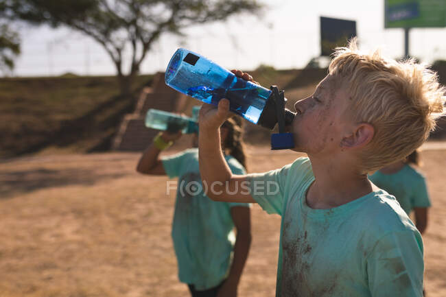 Un groupe de garçons et de filles caucasiens avec des t-shirts verts boueux et des visages sales se reposer et boire des bouteilles d'eau dans un camp de démarrage par une journée ensoleillée — Photo de stock