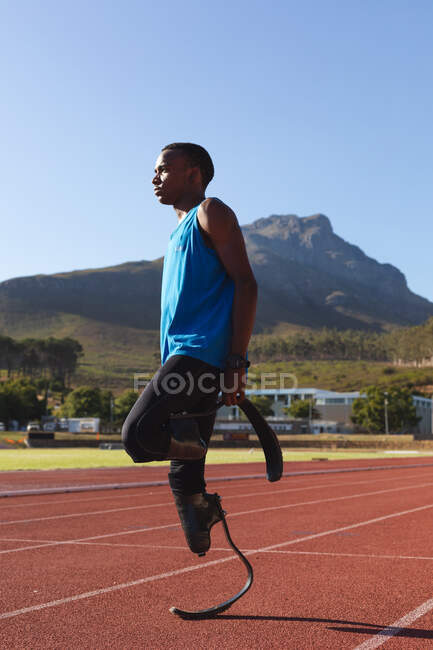 Fit, mixed race disabled male athlete at an outdoor sports stadium, preparing before workout stretching on race track wearing running blades. Disability athletics sport training. — Stock Photo