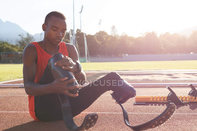 Fit, mixed race disabled male athlete at an outdoor sports stadium, sitting on race track adjusting running blades. Disability sport training. — Stock Photo