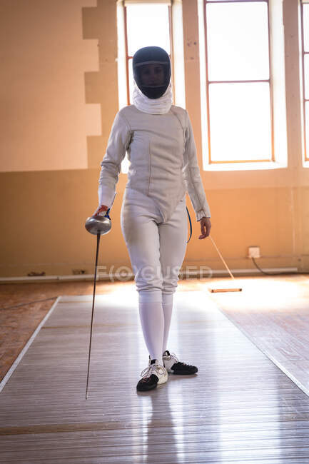 Портрет кавказской спортсменки, одетой в защитный костюм фехтовальщика во время тренировки по фехтованию, смотрящей в камеру и улыбающейся, держащей эпинефрин. Обучение фехтовальщиков в спортзале. — стоковое фото