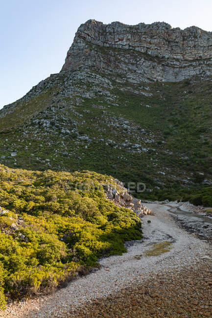 A walking path cutting through mountains with a tall rock face in the background in front of a blue sky on a sunny day. Beautiful natural scenery by the coast. — Stock Photo
