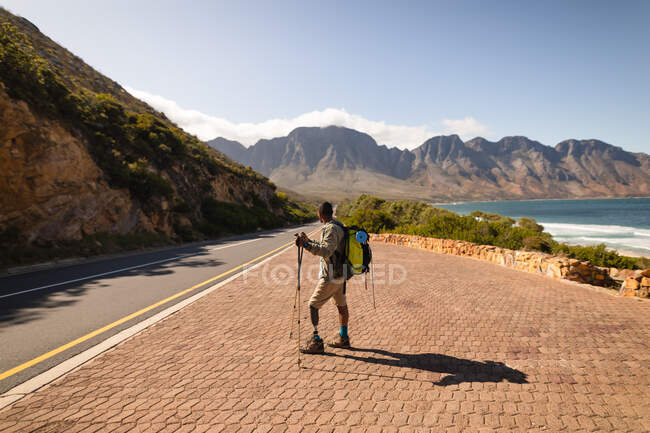 Athlète masculin de race mixte en forme et handicapé avec une jambe prothétique, profitant de son temps sur un voyage à la montagne, randonnée avec des bâtons, marche sur la route par la mer. Mode de vie actif avec handicap. — Photo de stock