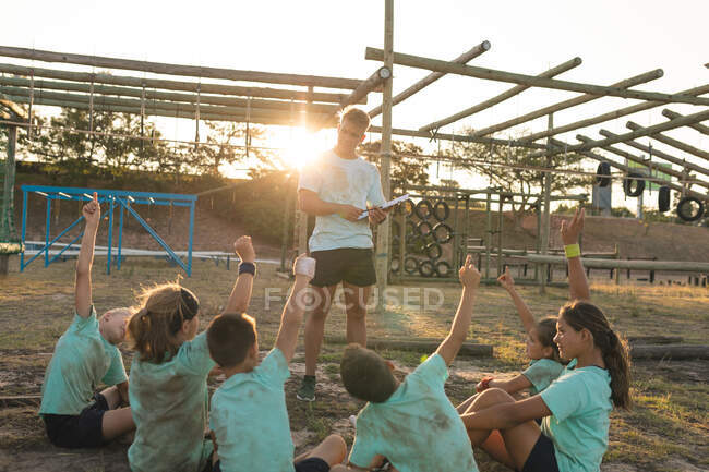 Группа кавказских мальчиков и девочек, слушающих инструкции кавказского тренера по фитнесу в лагере в солнечный день, сидящих на траве и поднимающих руки — стоковое фото