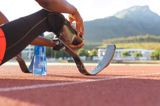 Low section of fit, disabled male athlete at an outdoor sports stadium, resting with water bottle on race track wearing running blades. Disability athletics sport training. — Stock Photo