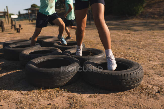 Low section of a group of kids at a boot camp on a sunny day, wearing green t shirts and black shorts, running through tyres on an obstacle course — Stock Photo