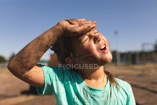 Tired, hot Caucasian girl at a boot camp on a sunny day, with mud on her face and arm and wearing a dirty green t shirt, wiping her brow — Stock Photo