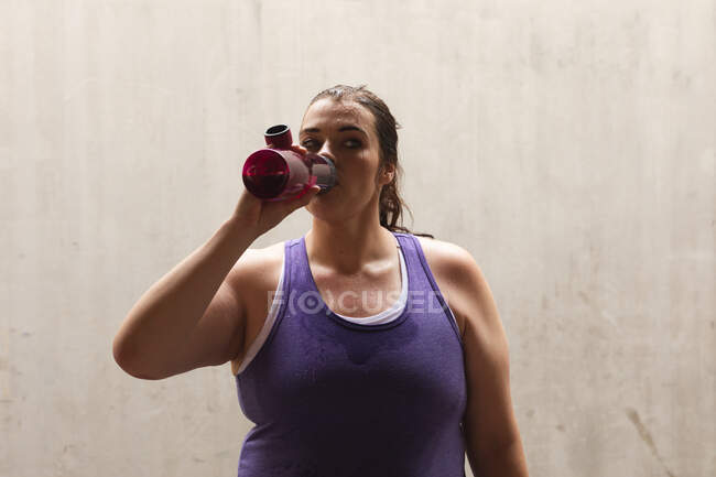 Кудрявая белая женщина с длинными темными волосами в спортивной одежде, занимающаяся спортом в городе, отдыхающая и пьющая из бутылки с водой во время тренировки — стоковое фото