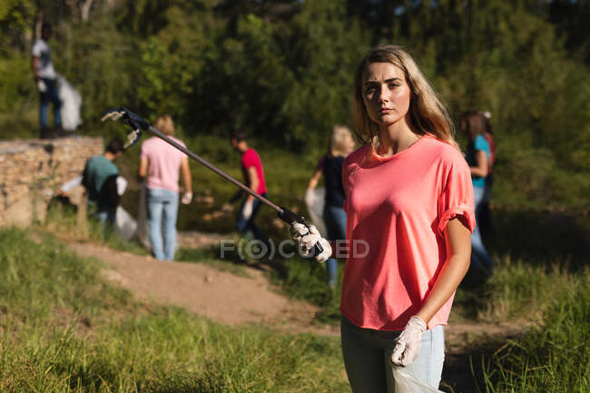 Portrait d'une femme caucasienne volontaire pour la conservation nettoyant la rivière à la campagne, ses amis ramassent des ordures en arrière-plan. Écologie et responsabilité sociale en milieu rural. — Photo de stock