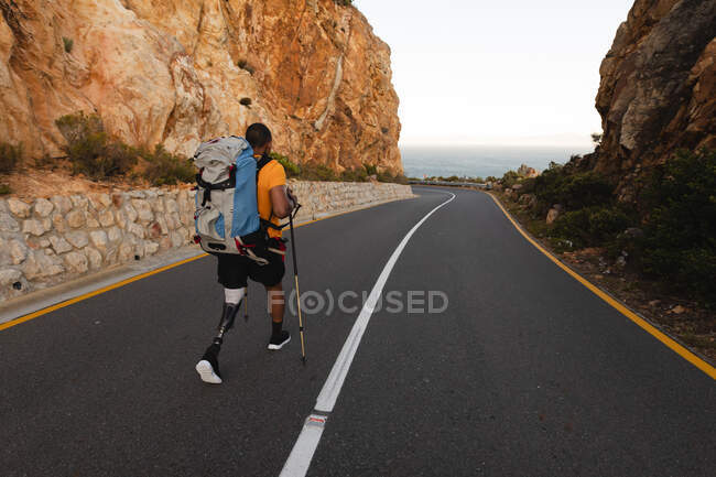 Athlète masculin en forme, handicapé, de race mixte avec une jambe prothétique, profitant de son temps sur un voyage à la montagne, randonnée, marche sur la route par la mer. Mode de vie actif avec handicap. — Photo de stock