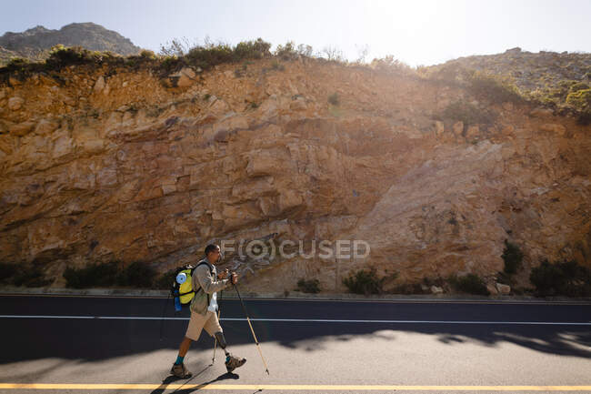 Athlète masculin en forme, handicapé, de race mixte avec une jambe prothétique, profitant de son temps sur un voyage à la montagne, randonnée avec des bâtons, marche sur la route dans les montagnes. Mode de vie actif avec handicap. — Photo de stock