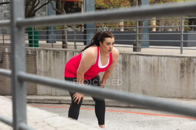 Кудрявая белая женщина с длинными темными волосами в спортивной одежде, тренирующаяся в городе, склоняющаяся вперед, чтобы отдохнуть во время тренировки, с современными зданиями на заднем плане — стоковое фото