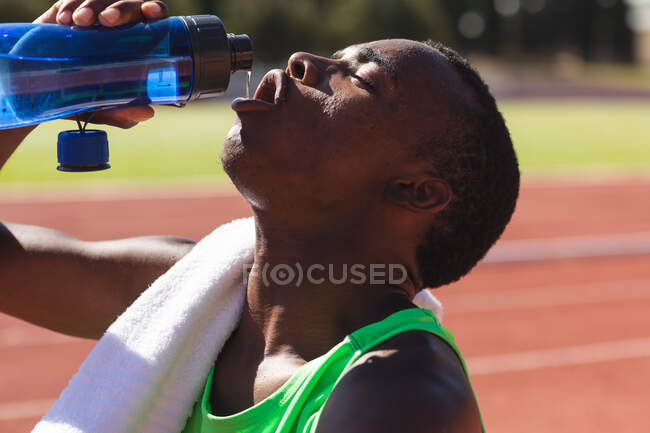 Fit, Mixed Race männlicher Athlet in einem Outdoor-Sportstadion, sitzt nach dem Rennen auf der Rennstrecke und trinkt Wasser mit einem Handtuch auf der Schulter. Leichtathletik-Training. — Stockfoto